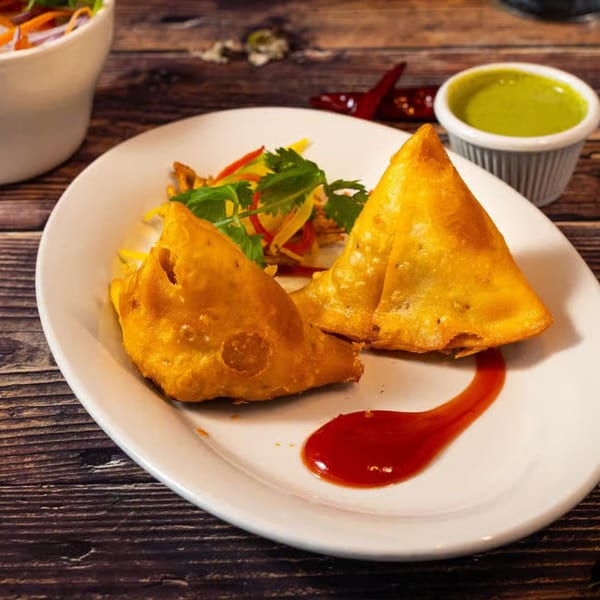 Two golden-brown samosas with a side of green chutney and red sauce on a white plate.