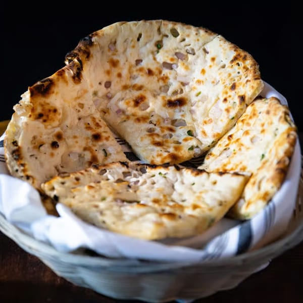 Basket of freshly baked Indian naan bread with charred spots, lined with a white and gray striped cloth.